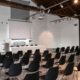 Corporate meeting room with rows of black chairs facing a stage and projector screen.