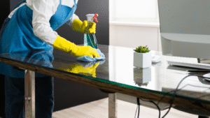 Cleaner disinfecting a glass office desk with gloves and spray bottle.