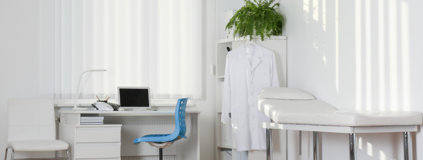 Bright medical exam room with clean desk, blue chair, white coat hanging, and exam table prepared for patients.