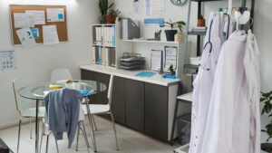 Healthcare staff room with workstations, medical coats, storage shelves, and organized surfaces in a clinical environment.